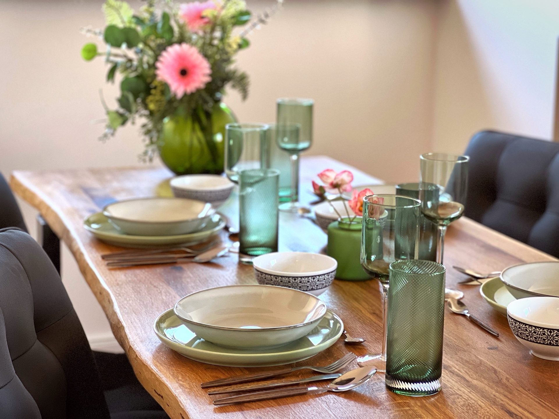 Gedeckter Holztisch mit grünen Gläsern, Tellern und Blumen